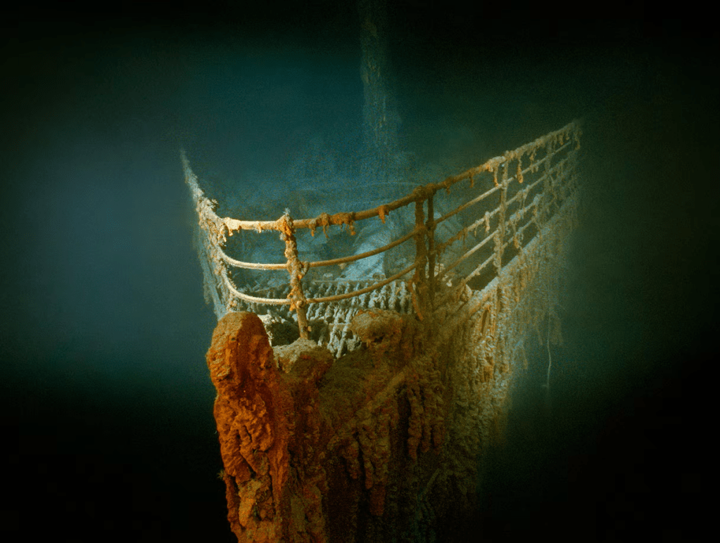 The bow section of the Titanic wreckage is partially covered in rust, surrounded by dark ocean water, showing remnants of its railings and decor.