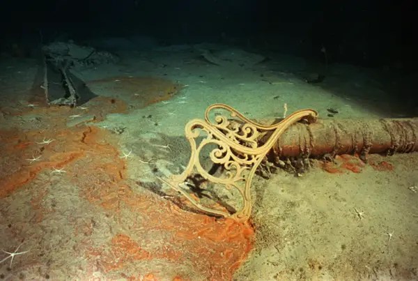 An ornate piece of furniture from the RMS Titanic wreckage resting on the ocean floor, surrounded by sediment and marine life. Here's one of those same benches at the bottom of the ocean